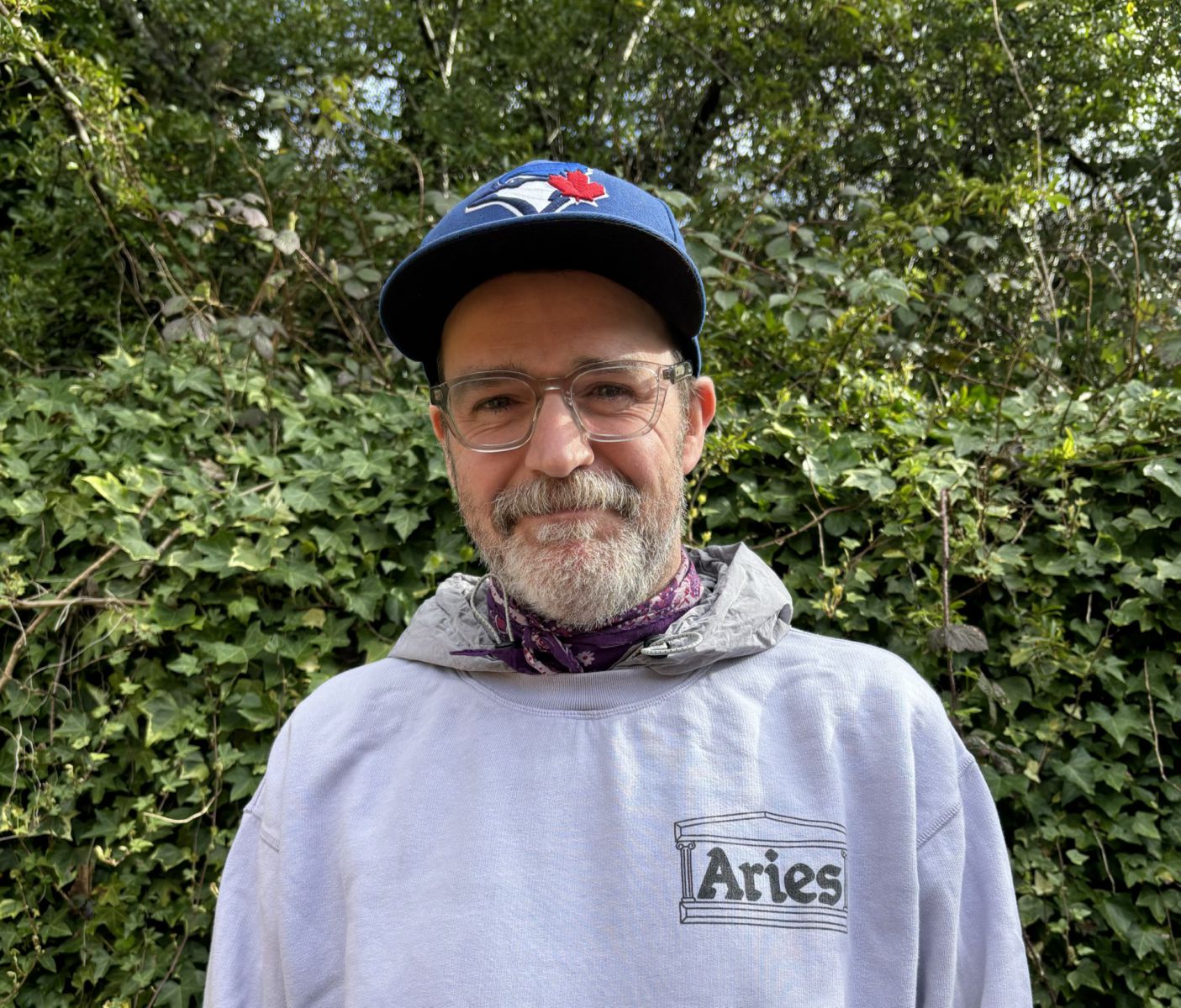 Dr Dave Lyons portrait. Dave stands in front of a green hedge. He wears a cap and glasses and a grey hoodie. He is white and has a beard.
